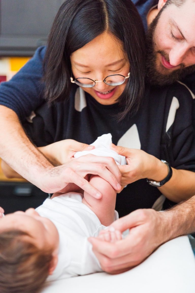 Une femme portant des lunettes et un homme barbu tiennent et regardent doucement un bébé habillé de blanc. Le bébé est allongé sur une surface blanche tandis que les deux adultes sourient, leur attention se concentrant tendrement sur le bébé. Cette image capture des moments précieux de famille, avec des touches d'éléments de cuisine dans l'arrière-plan flou.