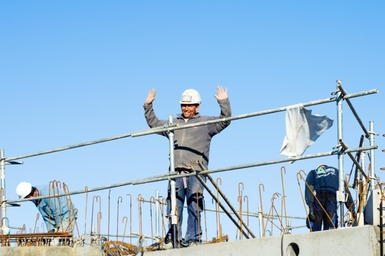 Un ouvrier du bâtiment portant un casque de sécurité blanc et un équipement de sécurité lève les deux bras en l'air en souriant. Il se tient debout sur une structure en béton avec des barres métalliques qui dépassent, entourée d'échafaudages. Un autre ouvrier portant un casque de sécurité blanc de Delta Construction à Bordeaux est penché en avant, travaillant sur le côté gauche de l'image. Le ciel est bleu clair.