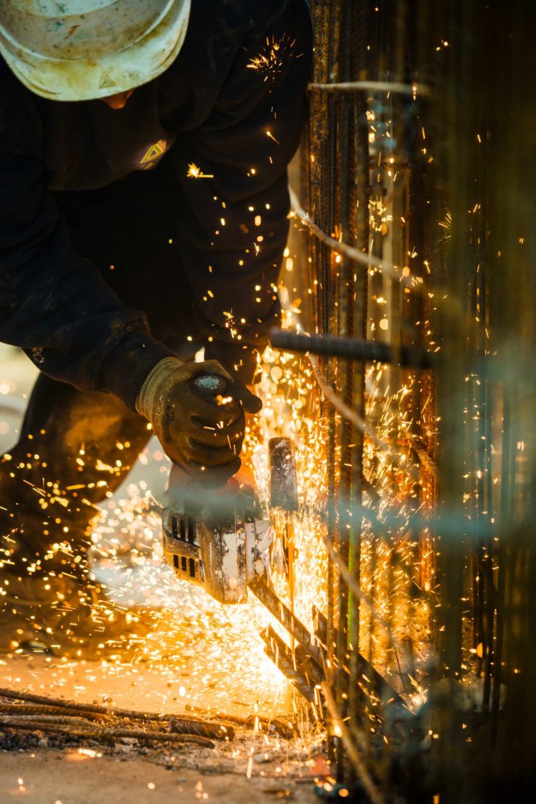 Un ouvrier de Delta Construction à Bordeaux, coiffé d'un casque et de gants, utilise une meuleuse d'angle pour découper des tiges métalliques. Des étincelles orange vif jaillissent de la surface, illuminant la zone. L'ouvrier est concentré sur sa tâche, les deux mains agrippées à l'outil. L'arrière-plan est flou, ce qui souligne l'intensité du travail.