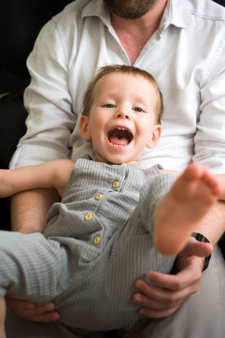 Un bambin en salopette grise à boutons en bois est assis sur les genoux d'un adulte. L'enfant a un grand sourire et rit, montrant son excitation et sa joie. L'adulte, vêtu d'une chemise blanche, tient l'enfant avec précaution, partiellement visible, le visage hors du cadre - un moment parfait pour tout photographe de famille bordelais à capturer.