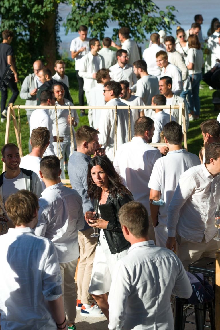 Une soirée conviviale se déroule en plein air par temps ensoleillé, avec une majorité d'hommes vêtus de chemises blanches et quelques femmes qui se mêlent. Une femme au premier plan porte une veste noire et tient un verre. Des arbres et de la verdure sont visibles à l'arrière-plan, ajoutant une touche dynamique à la scène de la soirée annuelle du Groupe Betclic.