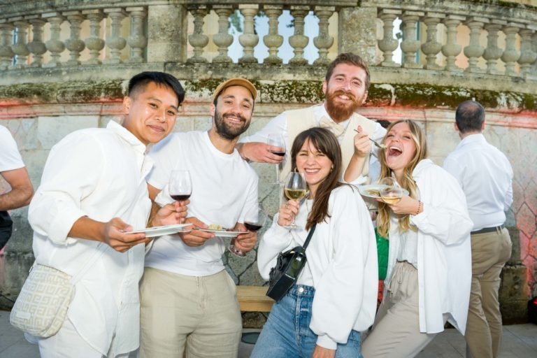 Un groupe de cinq personnes, trois hommes et deux femmes, se tiennent ensemble à l'extérieur lors de la Soirée annuelle du Groupe Betclic. Ils sourient et tiennent des verres à vin et des assiettes avec de la nourriture. Vêtus de vêtements décontractés de couleur claire, ils se tiennent près d'une balustrade en pierre ornée avec quelques autres participants en arrière-plan.