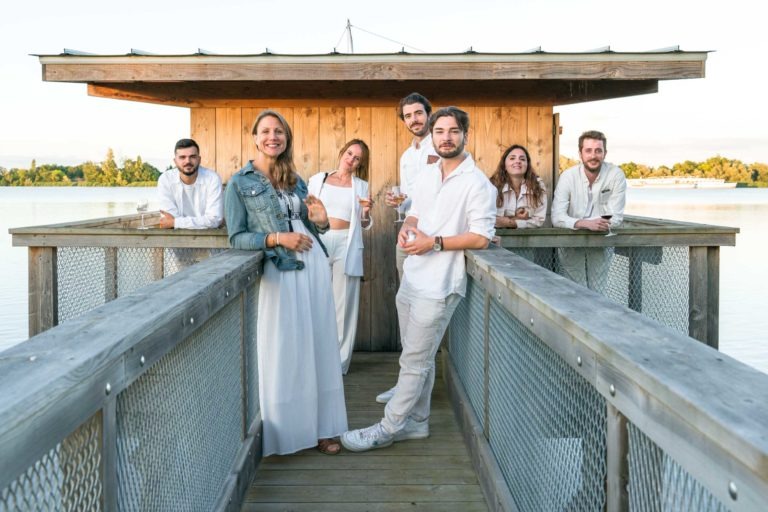 Un groupe de six personnes, trois hommes et trois femmes, se tiennent debout et s'assoient sur un quai en bois avec balustrades au bord d'un lac pendant la Soirée annuelle du Groupe Betclic. Ils sont détendus, vêtus de vêtements clairs et décontractés et sourient. Une structure en bois est en arrière-plan et des arbres sont visibles de l'autre côté de l'eau. Le ciel est partiellement nuageux.