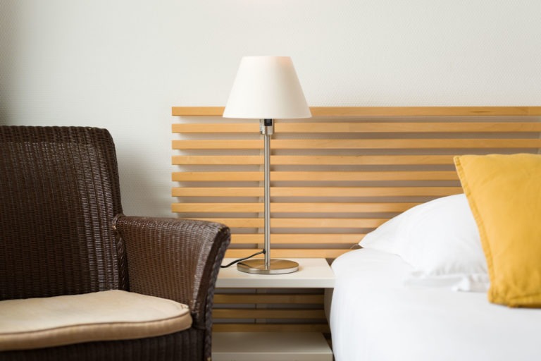 Une chambre moderne dans les Appartements De Vacances Bordeaux présente une chaise en osier avec un coussin beige clair à côté d'un lit avec des draps blancs et un oreiller jaune. Une table de chevet accueille une lampe en métal élégante avec un abat-jour blanc. La toile de fond est une tête de lit en bois avec des lattes horizontales contre un mur blanc uni.