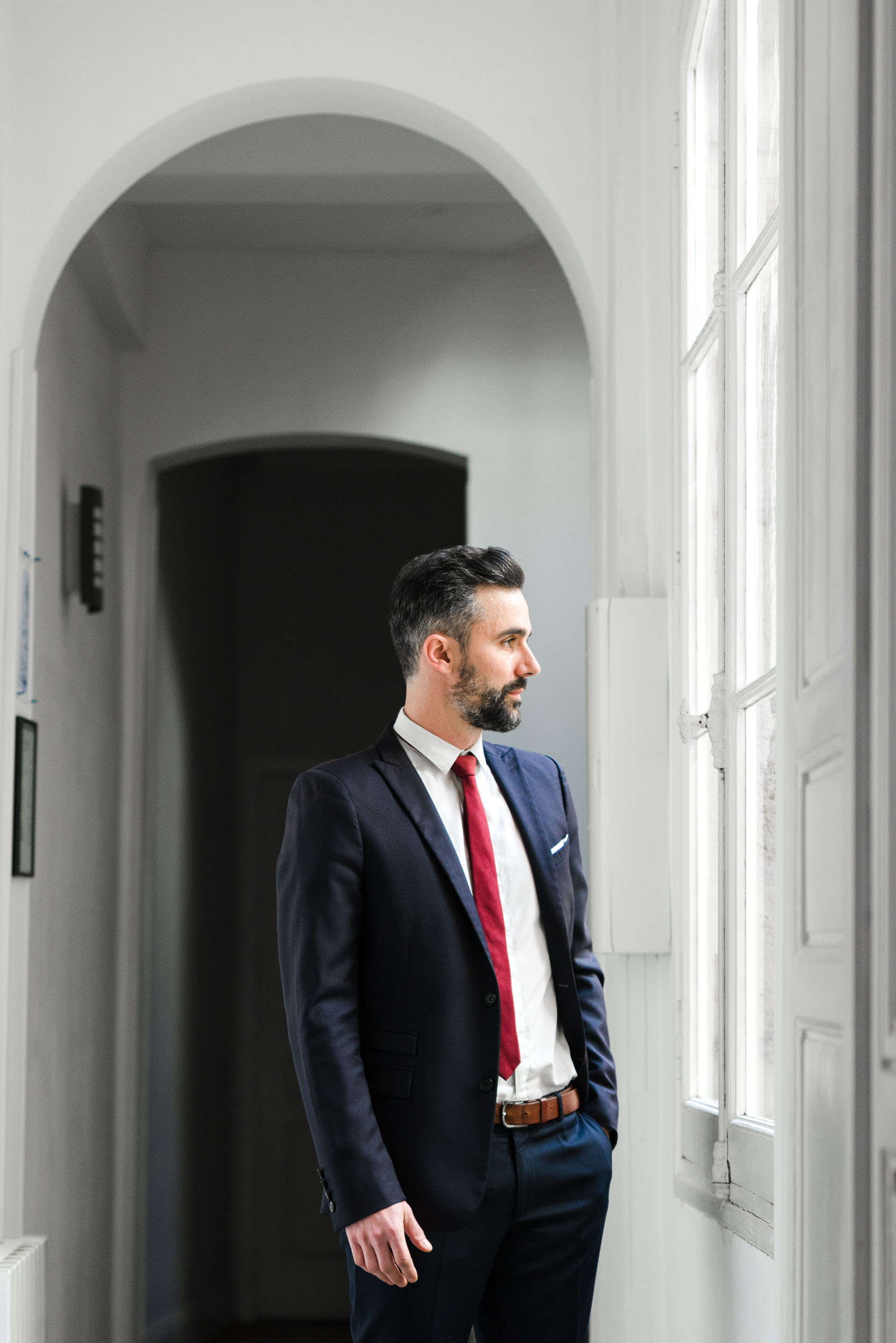 Un homme barbu, vêtu d'un costume sombre avec une cravate rouge, se tient debout dans un couloir très éclairé. Il regarde par une grande fenêtre à cadre blanc sur son côté droit. Le couloir présente des murs blancs et une porte cintrée en arrière-plan, idéale pour les portraits professionnels à Bordeaux.