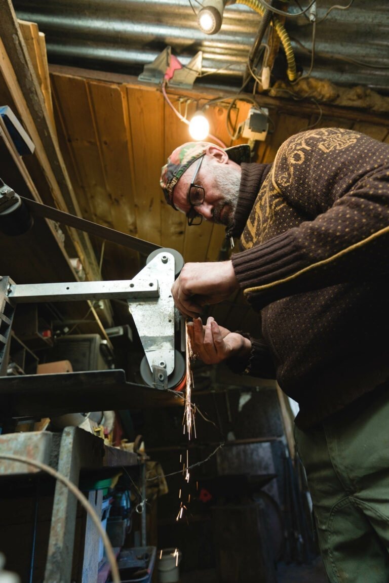 Un homme portant un pull marron et une casquette travaille intensément sur un outil mécanique dans un atelier faiblement éclairé. Il meule une pièce de métal, ce qui produit des étincelles. La pièce, remplie d'outils et d'objets éparpillés un peu partout, fait écho au savoir-faire du coutelier Philippe Sanchez, avec des murs en bois et des câbles apparents suspendus au plafond en tôle ondulée.