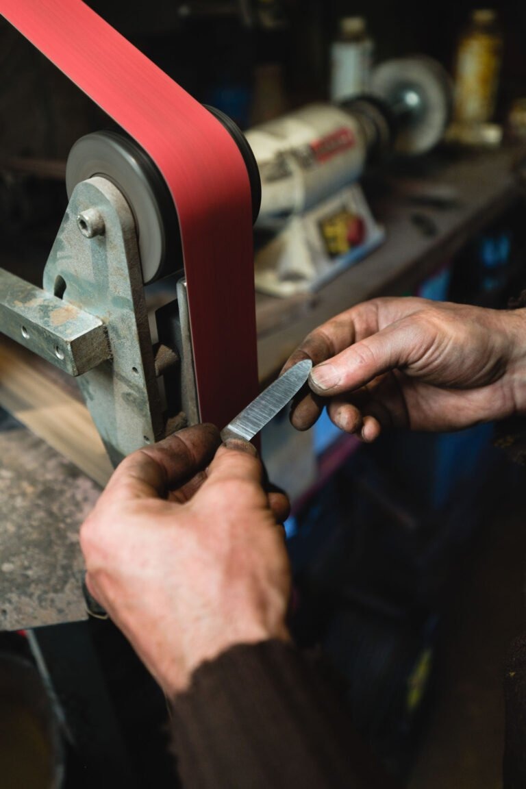 Gros plan sur les mains d'une personne tenant un objet métallique contre une ponceuse à bande rouge. Philippe Sanchez, coutelier de renom, façonne habilement la pièce métallique à l'aide de la bande abrasive rotative. La zone environnante comprend des machines et des outils d'établi flous, indiquant un environnement d'atelier.