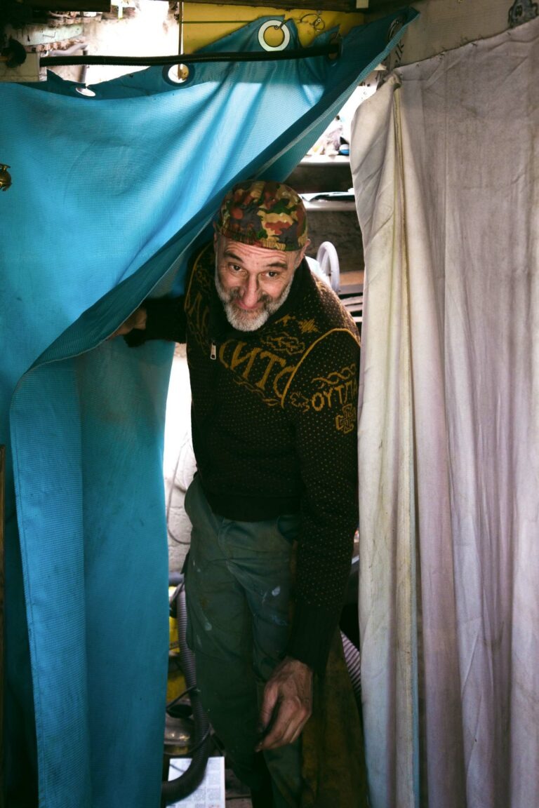 Un homme âgé avec une barbe, portant une casquette de camouflage, un pull à motifs sombres et un pantalon gris, tient un rideau turquoise à côté d'un rideau blanc. Il se tient dans un petit espace encombré avec divers objets partiellement visibles en arrière-plan. La scène de Philippe Sanchez coutelier capture un charme rustique.