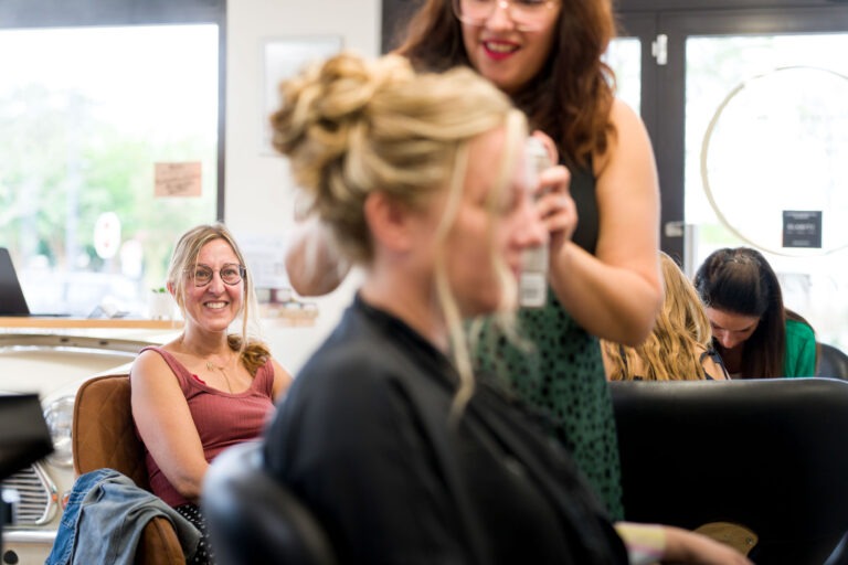 Une femme portant des lunettes et un débardeur rouge est assise et sourit sur une chaise de salon tandis qu'une autre femme, se préparant peut-être pour son mariage, se fait coiffer avec une cape marron drapée sur ses épaules. La styliste, tenant une bombe de laque, termine une coiffure complexe. D'autres clientes et stylistes sont visibles en arrière-plan.