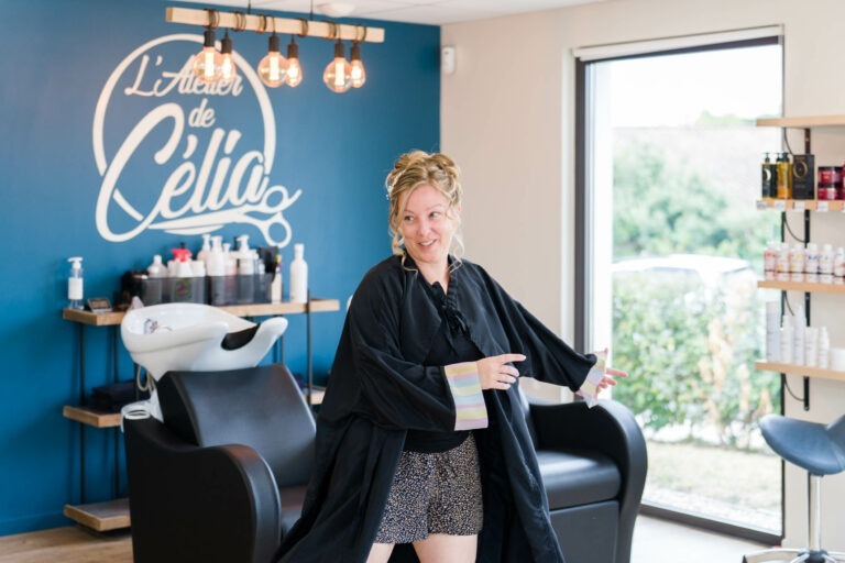 Une femme vêtue d'une robe de styliste noire sourit et pointe du doigt tout en se tenant dans un salon lumineux au décor moderne. Derrière elle, il y a des chaises de salon, des produits de soins capillaires et un mur bleu avec le texte « L'Atelier de Célia ». Des étagères avec d'autres produits sont visibles sur la droite, parfait pour votre mariage à Bordeaux.