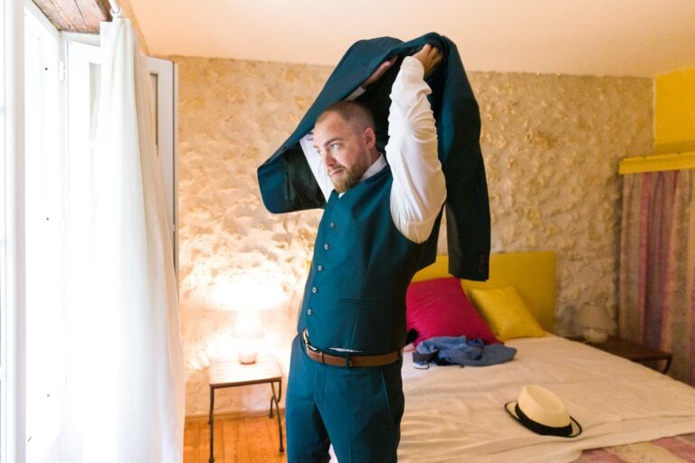 Un homme barbu se tient dans une chambre à coucher, enfilant une veste de costume vert foncé sur une chemise blanche. La pièce, qui rappelle le charme bordeaux avec ses murs en pierre, offre une scène chaleureuse avec des coussins colorés et un chapeau sur le lit. Une fenêtre avec des rideaux blancs et une petite lampe sur une table de nuit en bois complètent le décor.