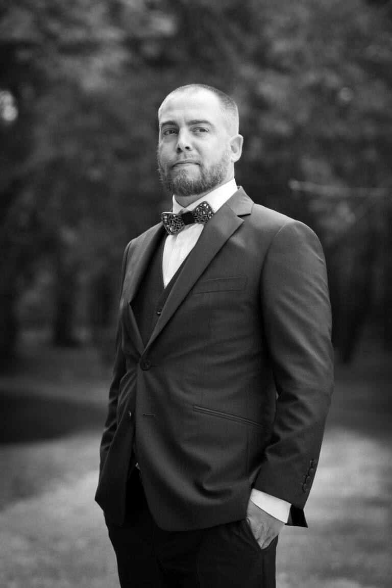 Un homme se tient debout dans une zone boisée, vêtu d'un costume sombre avec un nœud papillon et une barbe bien taillée. Une main est dans sa poche et il regarde l'objectif avec assurance. L'image, qui rappelle les portraits de mariage d'époque, est en noir et blanc, avec l'arrière-plan légèrement flou pour mettre en valeur le sujet.