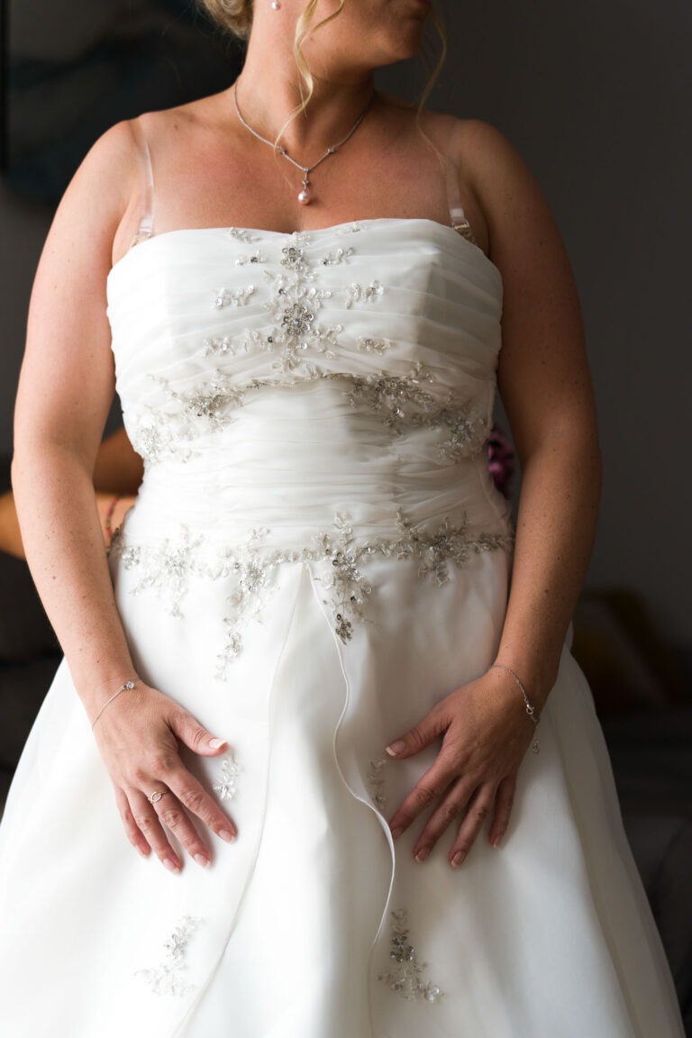 Une femme porte une robe de mariée blanche ornée de perles et de broderies florales. Elle a les mains posées sur les côtés, mettant en valeur son délicat bracelet. Ses cheveux sont relevés et elle porte un collier. L'arrière-plan, rappelant les vignobles de Bordeaux, est légèrement flou, mettant en valeur sa robe.