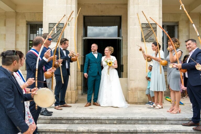 Une mariée en robe blanche et un marié en costume bleu canard, se tenant la main et portant des fleurs, se tiennent à l'entrée d'un bâtiment. Huit capoeiristes forment une arche avec des arcs musicaux au-dessus du couple. L'ambiance est festive, les participants étant en tenue de soirée. Des colonnes de pierre encadrent l'entrée à Bordeaux.