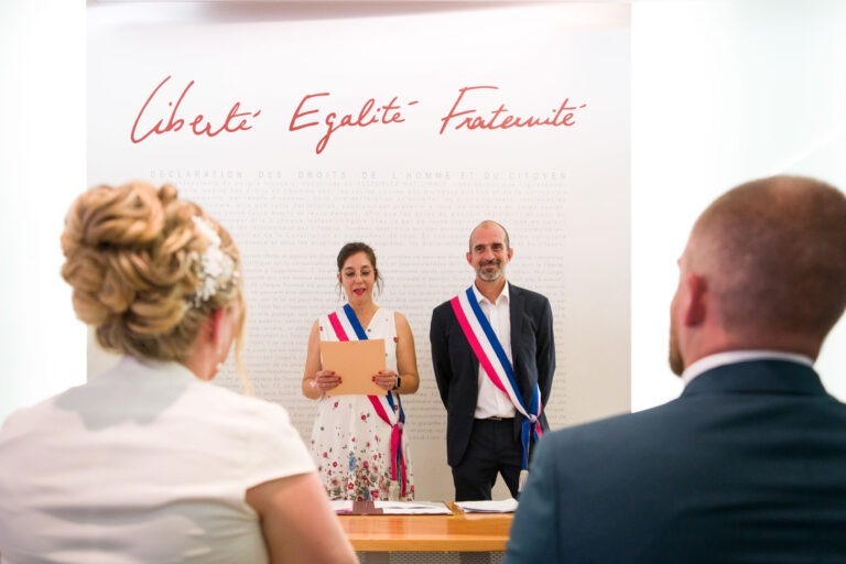 Une cérémonie de mariage se déroule devant un mur sur lequel est inscrit « Liberté, Egalité, Fraternité ». Deux officiants portant l'écharpe tricolore française se tiennent face au couple, la femme tenant un papier. Le couple est assis, les cheveux de la mariée relevés et le marié en costume bordeaux.