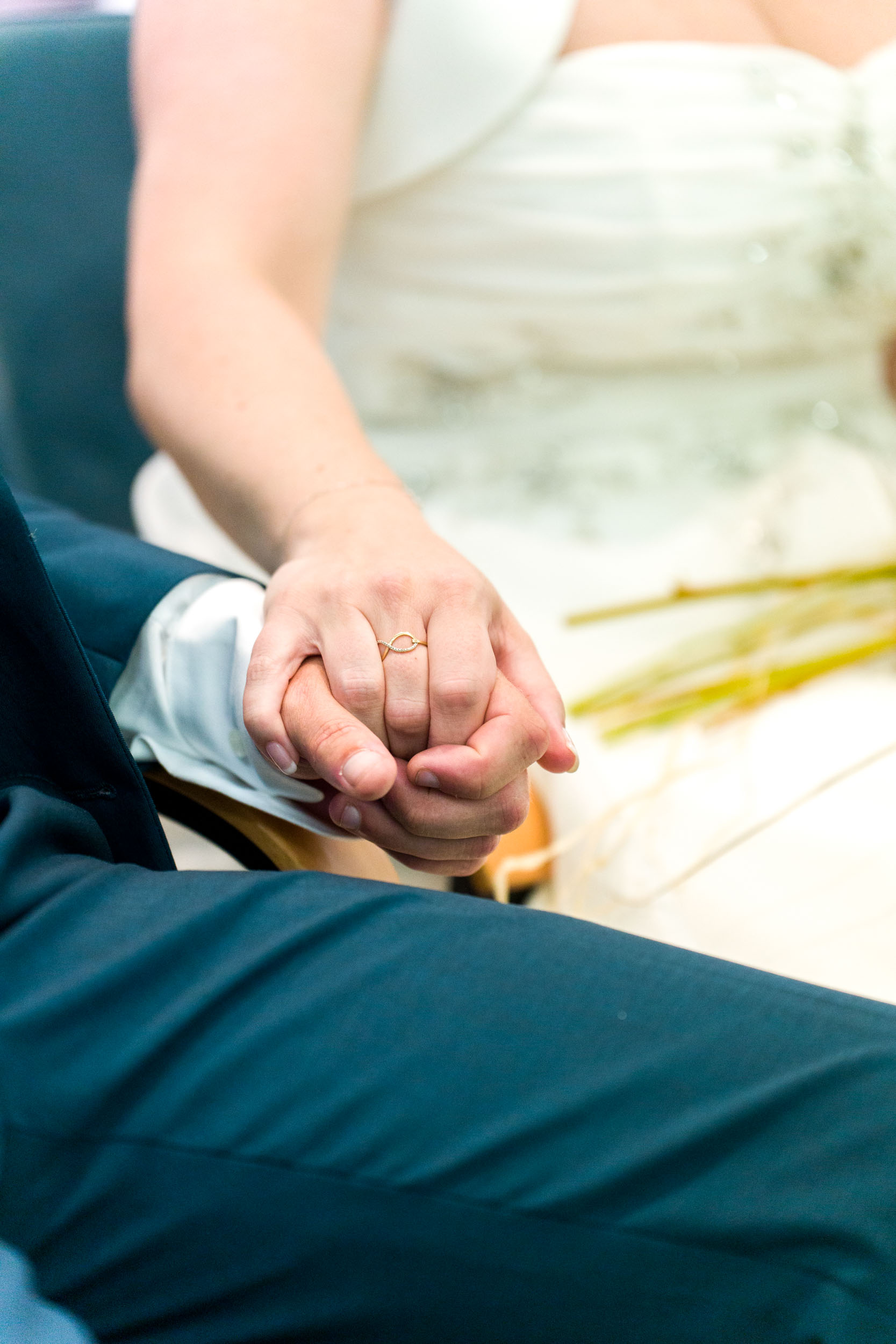 Gros plan d'une mariée et d'un marié se tenant la main. La main de la mariée, ornée d'une délicate bague, repose sur celle du marié. La mariée est vêtue d'une robe blanche avec des broderies florales visibles, tandis que le marié porte un costume sombre. Ce moment intime capture l'essence de leur mariage et de leur lien émotionnel profond.