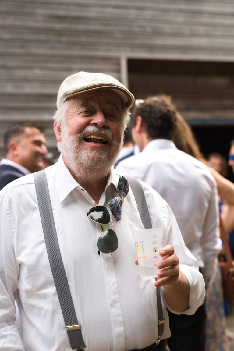 Un homme âgé avec une barbe blanche rit joyeusement lors d'un rassemblement en plein air, peut-être un mariage. Il porte une chemise blanche, des bretelles grises, une casquette plate de couleur claire, un nœud papillon et des lunettes de soleil suspendues à sa chemise. Il tient un gobelet en plastique transparent. Des personnes et une structure en bois sont visibles dans l'arrière-plan flou.