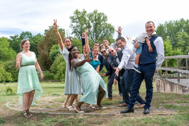 Un groupe de personnes joyeuses, habillées de manière formelle ou semi-formelle, posent avec énergie pour une photo en plein air lors de ce qui semble être un mariage festif à Bordeaux. Certains font des signes de paix et rient, tandis qu'un homme tient un enfant dans ses bras. Ils sont entourés d'une végétation luxuriante et d'une clôture en bois, renforçant l'atmosphère de fête.