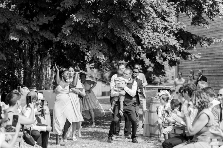 Une image en noir et blanc montre une joyeuse cérémonie de mariage en plein air à Bordeaux. Une foule est assise de chaque côté d'une allée. Un homme en costume et cravate porte joyeusement un jeune garçon dans l'allée. Plusieurs adultes le suivent et les invités applaudissent, immortalisant le moment sur leurs téléphones. Des arbres et un bâtiment sont en arrière-plan.