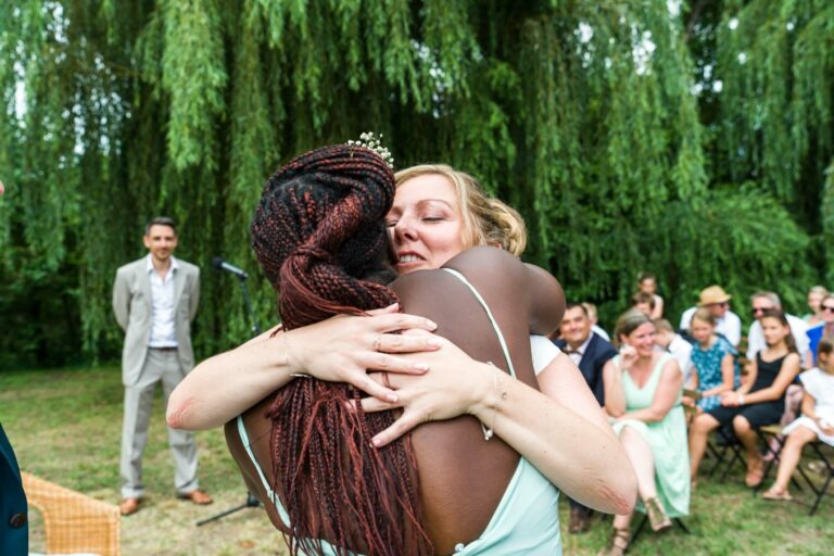 Deux femmes se serrent dans leurs bras, l'une avec des cheveux tressés et un petit diadème, l'autre avec des cheveux blonds. Elles se tiennent dehors devant un groupe de personnes assises, entourées d'arbres verts et d'herbe luxuriante. Un homme en costume les observe de loin, ajoutant à l'atmosphère festive de ce qui ressemble à un mariage à Bordeaux.