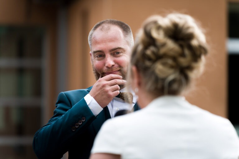 Un homme barbu et aux cheveux courts, vêtu d'un costume bleu canard, sourit et se tient le menton avec la main. Il regarde une femme aux cheveux blonds attachés en un élégant chignon, portant un haut ou une robe blanche. L'arrière-plan est flou mais suggère un décor extérieur à Bordeaux.