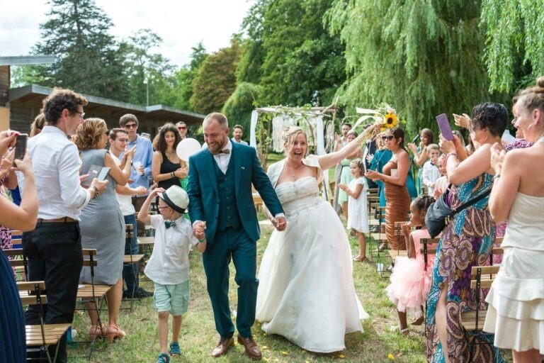 Une mariée et un marié marchent main dans la main dans une allée extérieure, souriant joyeusement. La mariée agite un tournesol et le marié tient la main d'un garçon portant un chapeau. Les invités de chaque côté applaudissent et acclament, certains prenant des photos. Des arbres et une arche décorée peuvent être vus en arrière-plan, ce qui rend la scène du mariage festive et animée.