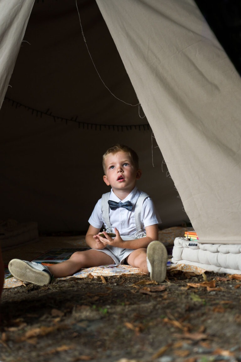 Un jeune garçon est assis dans une grande tente beige lors d'un mariage, vêtu d'une chemise blanche et d'un nœud papillon foncé. Il regarde vers le haut avec une expression curieuse, tenant un petit objet dans ses mains. Autour de lui se trouvent quelques livres éparpillés et un tapis rembourré sur le sol. Les volets de la tente sont ouverts, permettant à la lumière d'entrer.