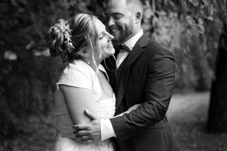 Une photo en noir et blanc d'un couple le jour de leur mariage montre la mariée, vêtue d'une robe blanche et d'un postiche fleuri, souriante, les yeux fermés. Le marié, habillé d'un costume et d'un nœud papillon, l'embrasse d'un air joyeux au milieu des arbres. Ce moment enchanteur met en valeur leur mariage à Bordeaux.