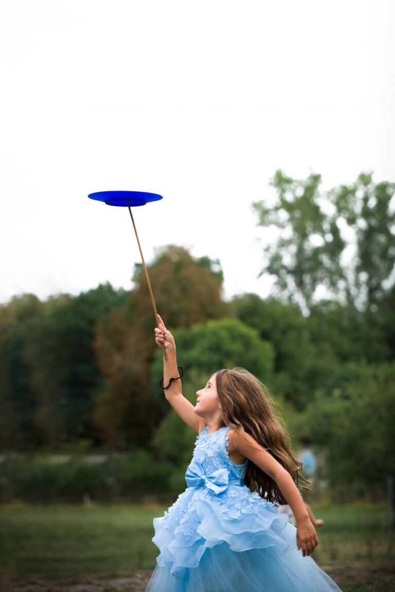 Une jeune fille vêtue d'une robe bleu clair à volants tient en équilibre une assiette bleue sur un bâton. Elle est dehors, dans une zone herbeuse avec des arbres en arrière-plan, les yeux levés vers l'assiette avec un bras tendu. Ses cheveux bruns flottent librement alors qu'elle se concentre sur l'équilibre de l'assiette, évoquant la grâce des capoeiristes en mouvement.