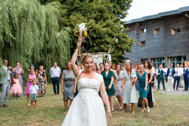 Une mariée en robe blanche se tient au premier plan, souriante, tout en tenant un bouquet. Derrière elle, un groupe de femmes attendent avec impatience, se préparant à attraper le bouquet. Le décor extérieur de Bordeaux comprend une végétation luxuriante et un bâtiment au bardage en bois. Divers invités observent en arrière-plan.