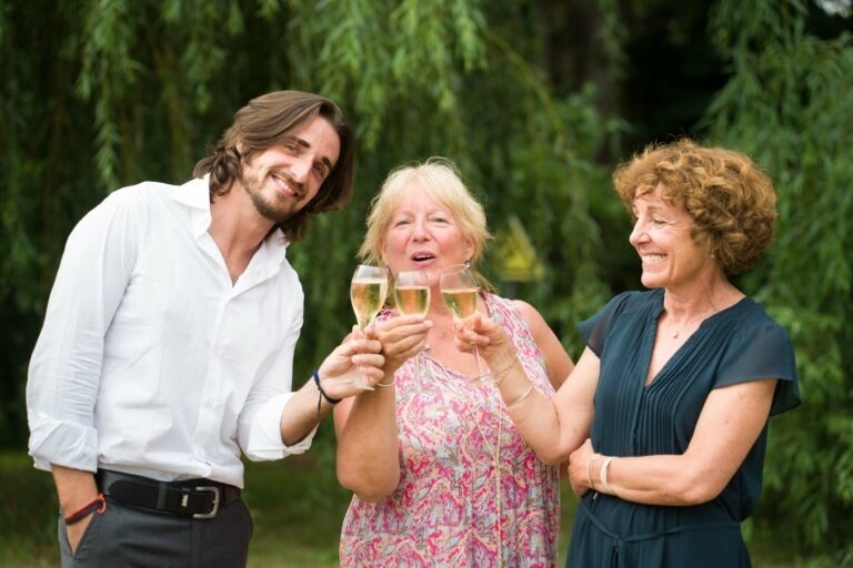 Trois personnes se tiennent à l'extérieur, levant des verres de champagne et souriant. Un homme portant une chemise blanche aux manches retroussées est à gauche, tandis que deux femmes sont à droite. La femme du milieu tient un verre à deux mains, porte une robe colorée suggérant des teintes bordeaux vibrantes, et la femme de droite porte une robe sombre. De la verdure est à l'arrière-plan.