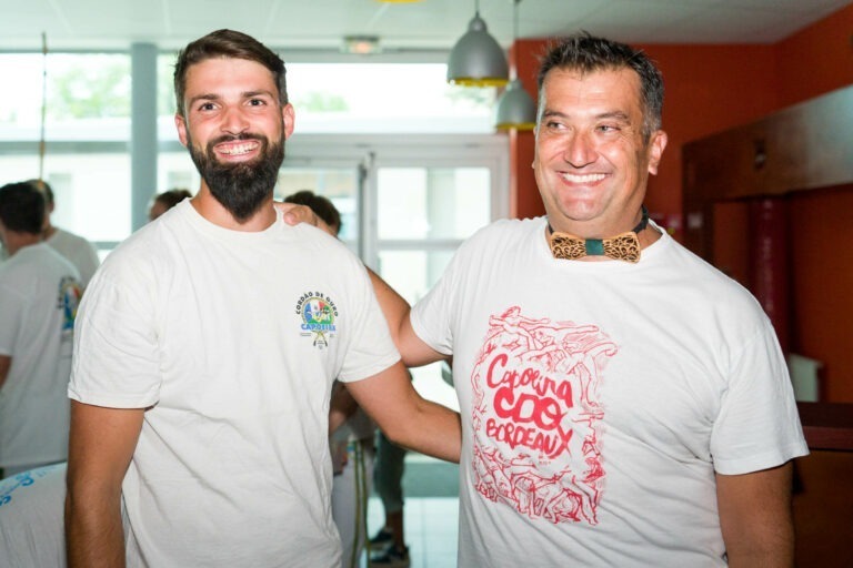 Deux hommes souriant côte à côte. L'homme de gauche, probablement un capoeiriste, a une barbe et porte un T-shirt blanc avec un logo sur la manche. L'homme de droite a les cheveux gris, porte un T-shirt blanc avec "Coupe de France" en rouge et un nœud papillon en bois. Ils sont à l'intérieur avec des lumières colorées en arrière-plan.