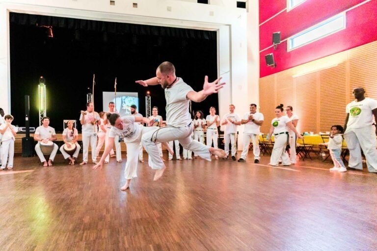 Deux hommes exécutent un mouvement de capoeira dynamique dans une salle lumineuse au sol en bois. L'un d'eux est en l'air, en plein coup de pied, tandis que l'autre esquive. Tous deux portent des tenues blanches avec des logos de capoeira. Des participants enthousiastes et un public en uniformes de capoeira blancs les entourent, applaudissent et acclament, un peu comme on le verrait lors d'un mariage de capoeiristes à Bordeaux.