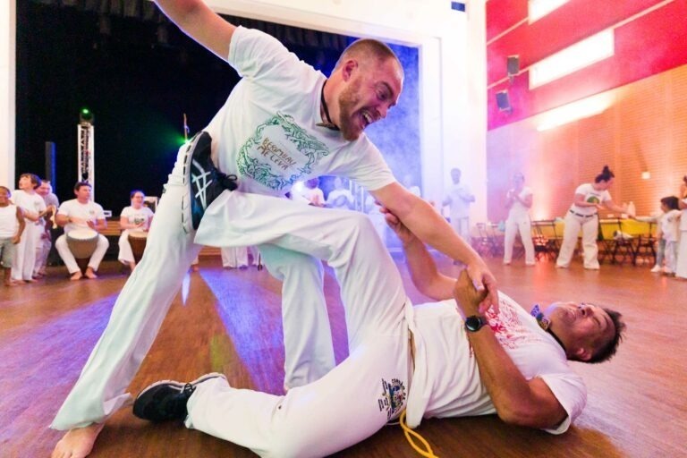 Deux hommes en tenue blanche d'arts martiaux pratiquent la capoeira dans une salle lumineuse au sol en bois. L'un des deux est au sol, tenant le bras de l'homme debout, tandis que l'homme debout se penche vers lui, saisissant l'épaule de l'homme au sol. En arrière-plan, d'autres capoeiristes bordelais se joignent à eux avec des instruments de musique.