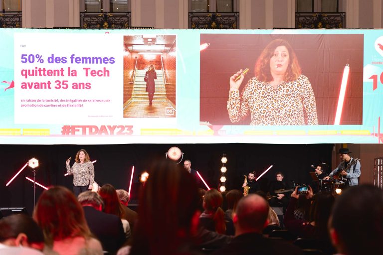 Une femme s'exprime sur scène lors de la French Tech Night 2023, avec "50% des femmes quittent la Tech avant 35 ans" affiché sur un grand écran derrière elle. Le public est assis, et des musiciens jouent d'un instrument à droite. Le hashtag de l'événement "#FTDAY23" est visible en arrière-plan.