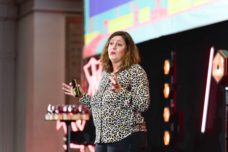 Une femme aux cheveux châtains mi-longs, vêtue d'un chemisier à imprimé léopard et d'un jean foncé, s'exprime sur scène lors de la French Tech Night 2023. Elle tient une petite télécommande dans une main et fait des gestes avec l'autre. L'arrière-plan présente un décor illuminé avec des lumières et un écran coloré affichant des graphiques.