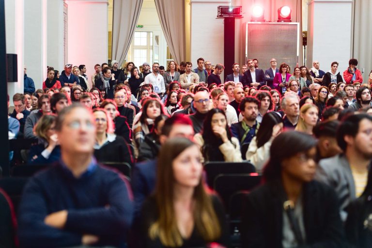 Un large public est assis et debout dans une salle bien éclairée, le regard tourné vers l’avant. En arrière-plan, un grand écran et des lumières projettent une teinte rouge. Les participants, vêtus de tenues décontractées ou business-casual, reflètent l’ambiance formelle de la French Tech Night 2023.