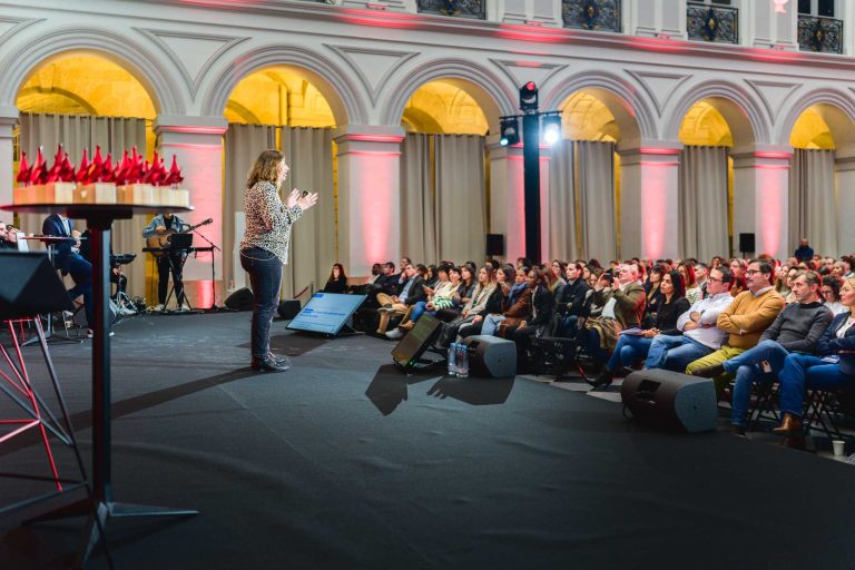 Un orateur se tient sur scène devant un public assis dans une grande salle élégante à l'architecture voûtée et à l'éclairage tamisé lors de la French Tech Night 2023. Le public, attentif, est disposé en rangées. Du matériel de musique et une guitare sont visibles sur le côté gauche de la scène.