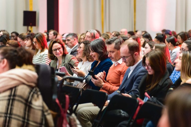 Un groupe diversifié de personnes est assis sur des rangées de chaises, en train d'interagir avec leurs téléphones et entre eux. Les participants, vêtus de tenues décontractées et professionnelles, semblent être à la French Tech Night 2023. L'arrière-plan est doucement éclairé par un éclairage rouge, créant une ambiance chaleureuse.