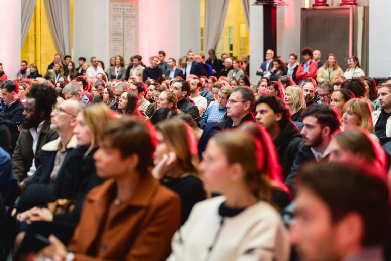 Un large groupe de personnes assises écoutent attentivement dans une salle spacieuse et bien éclairée. Le public de la French Tech Night 2023 est très diversifié et comprend des personnes d'âges et de sexes variés. Certains sont tournés vers l'avant, tandis que d'autres se tournent légèrement sur le côté. L'éclairage rouge crée une ambiance chaleureuse dans la salle.