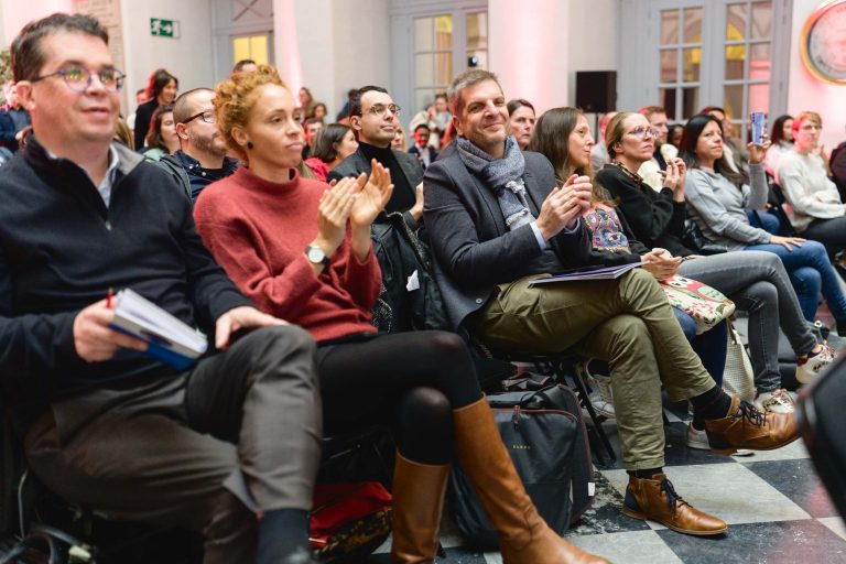 Un groupe diversifié de personnes est assis sur des chaises dans une salle bien éclairée avec de hautes fenêtres, écoutant attentivement une présentation à la French Tech Night 2023. Certains individus applaudissent, tiennent des cahiers ou des stylos. Le sol est orné d'un motif à carreaux noir et blanc, et l'atmosphère semble engageante et positive.