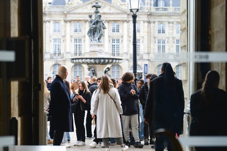 Un groupe de personnes se tient sous une arcade, donnant sur une place avec une grande fontaine et un bâtiment historique décoré en arrière-plan. La foule semble engagée dans diverses activités, avec des individus discutant et d'autres regardant leurs téléphones, vérifiant peut-être les mises à jour pour la French Tech Night 2023. L'atmosphère est vibrante et animée.