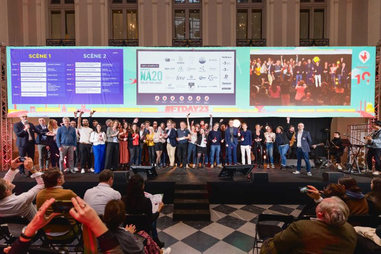 Lors de la French Tech Night 2023, un grand groupe de personnes se tient sur une scène dans une salle décorée avec de hauts plafonds et de hautes fenêtres. Deux grands écrans affichent des informations derrière eux, notamment les horaires et les logos des sponsors. Les membres du public sont assis devant, certains avec les mains levées ou prenant des photos. Diverses lumières illuminent la scène.