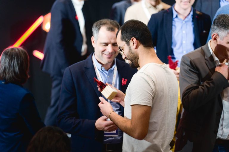 Lors de la French Tech Night 2023, un homme en t-shirt blanc tient et montre une boîte en bois à un homme en costume. Ils font partie d'un groupe plus large de personnes qui interagissent de manière décontractée et sourient. L'arrière-plan affiche des individus flous et des traînées de lumière rouge, créant une atmosphère informelle et conviviale.