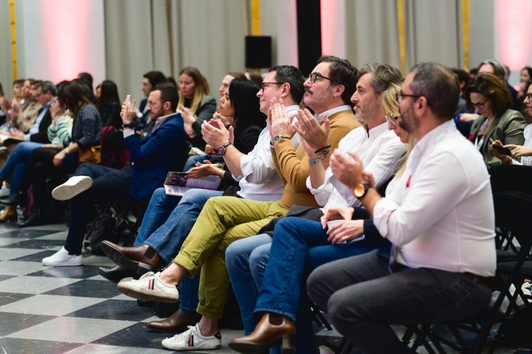 Un groupe de personnes assises en rangées de chaises, regardant attentivement vers l'avant lors de la French Tech Night 2023. Certaines personnes applaudissent, montrant leur engagement et leur appréciation. Le décor est intérieur avec un sol à carreaux et un arrière-plan doucement éclairé par des rideaux drapés.