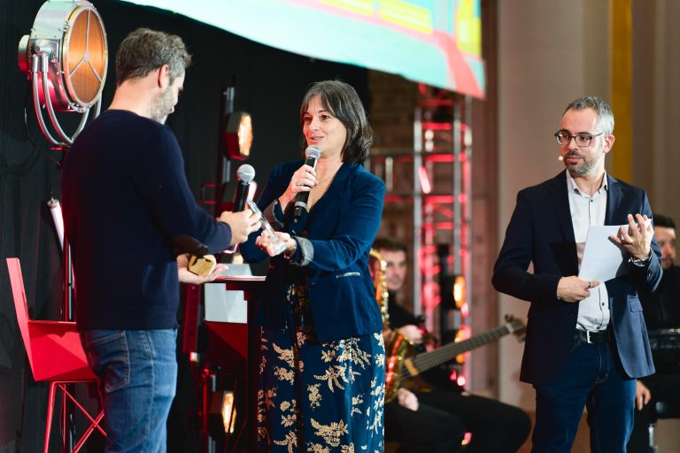 Lors de la French Tech Night 2023, trois personnes sont sur scène. Un homme à gauche, en chemise sombre, tend un micro à une femme au milieu, vêtue d'une tenue sombre à fleurs et tenant un autre micro. Un homme à droite, vêtu d'un costume sombre avec une chemise blanche, tient des papiers et observe.