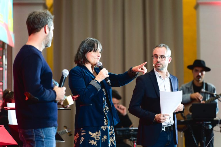 Trois personnes se tiennent sur une scène de la French Tech Night 2023. Une femme au centre parle dans un micro tout en pointant du doigt. Elle est entourée de deux hommes, l'un tenant un micro et l'autre des papiers. En arrière-plan, un musicien coiffé d'un chapeau joue du clavier électronique. Le décor semble formel.