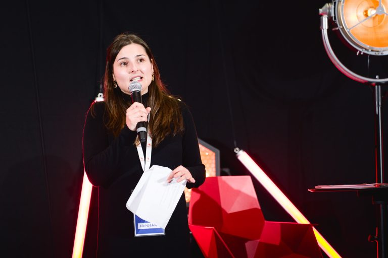 Une personne aux cheveux longs et noirs se tient sur une scène de la French Tech Night 2023, tenant un micro dans une main et des papiers dans l'autre. Elle porte un haut noir et un badge nominatif, et a l'air sérieux lorsqu'elle parle. En arrière-plan, des formes géométriques rouges et des néons attirent le grand projecteur à droite.