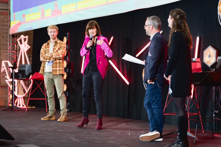 Quatre personnes se tiennent sur une scène avec un fond noir et quelques néons pendant la French Tech Night 2023. Une femme en veste rose parle dans un micro. À sa droite, un homme en blazer tient un papier et une autre femme sourit. À sa gauche, un homme en chemise à carreaux les regarde. Un écran aux graphismes colorés se trouve au-dessus d'eux.