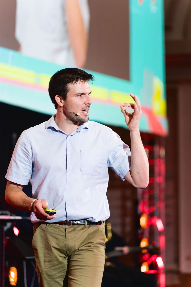 Un homme portant une chemise bleu clair et un pantalon vert fait une présentation à la French Tech Night 2023. Il tient un petit appareil dans sa main droite et fait un geste « OK » avec sa main gauche. Il porte un micro-casque et un grand écran avec des graphiques colorés est visible en arrière-plan.